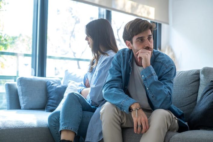 Young millennial couple sitting back to back on a sofa at home, sad and upset, ignoring each other, struggling with rela